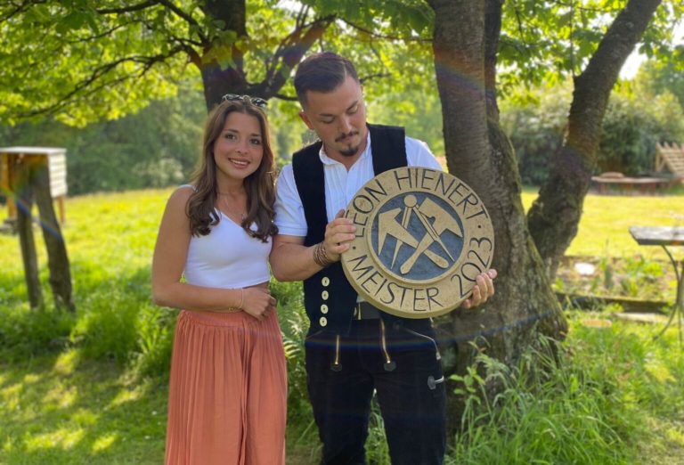 Inhaber von Firstetage, Leon Hienert mit Frau, mit Holzplakette für seinen Beruf als Dachdeckermeister.